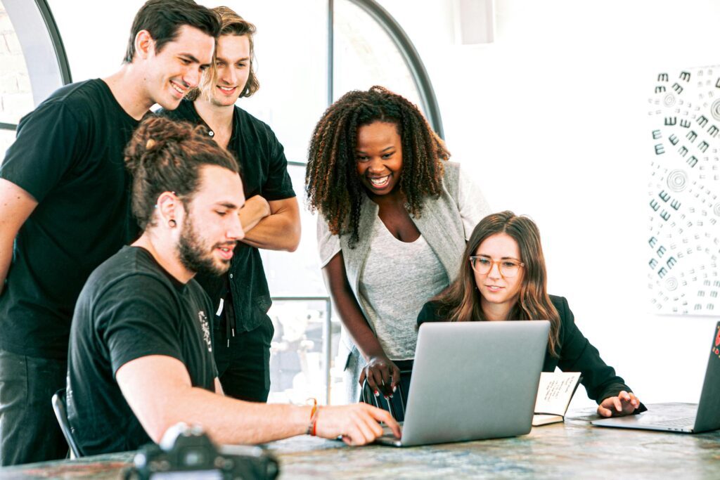 A diverse group of young professionals collaborate and share ideas around a laptop in a bright, modern office